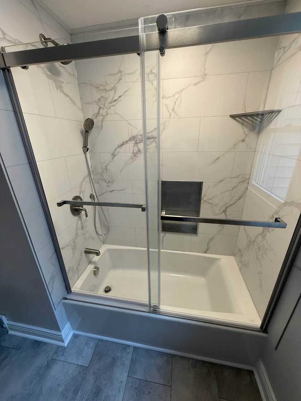 Bathtub with veined wall tiles, a recessed shower niche, and sliding glass doors.